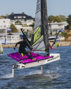 En talentfull sjømann manøvrerer en Waszp hydrofoil-seilbåt gjennom hakkete vann nær et kystområde, og viser frem bemerkelsesverdige sportsferdigheter med hus synlige i bakgrunnen.