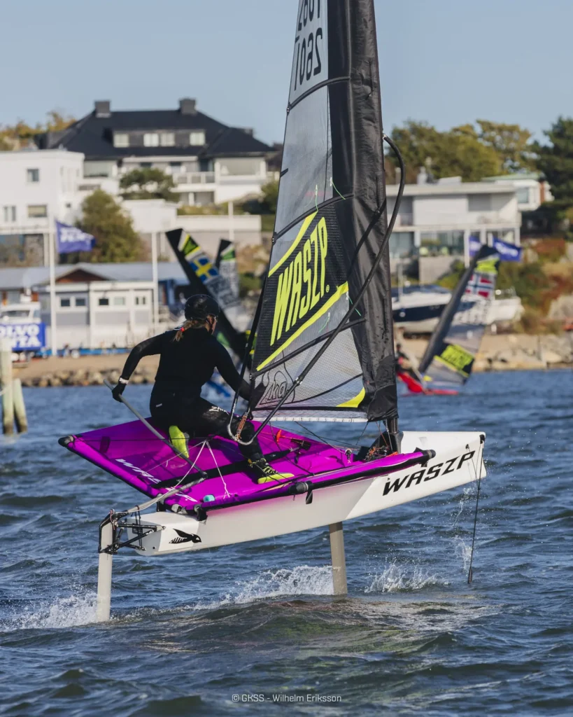 En talentfull sjømann manøvrerer en Waszp hydrofoil-seilbåt gjennom hakkete vann nær et kystområde, og viser frem bemerkelsesverdige sportsferdigheter med hus synlige i bakgrunnen.
