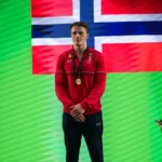 En utøver med gullmedalje står foran et norsk flagg mot grønn bakgrunn.