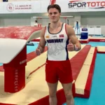 En mannlig gymnast i norsk uniform står i en gymnastikkarena og gir to tomler opp, med hopputstyr synlig i bakgrunnen.